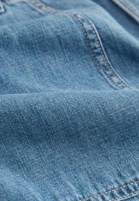 Denim tkanina u svijetloplavoj boji s blagom teksturom, koja prikazuje suptilno horizontalno tkanje i narančaste šavove duž rubova.
