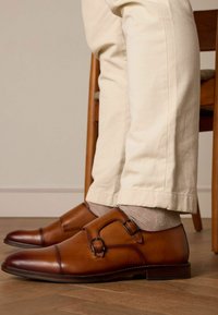 Chaussures en cuir marron à double boucle avec fermetures, texture lisse, bout pointu, associées à un pantalon couleur crème et des chaussettes beige.