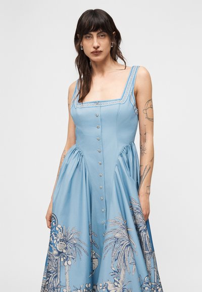 Γυναίκα με μακριά σκούρα μαλλιά και τατουάζ φορά αμάνικο μπλε φόρεμα με κουμπιά μπροστά και τύπωμα φοίνικα στη φούστα.