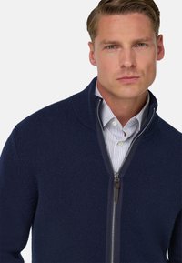 Marinblå zip-up tröja i strukturerad tyg med hög krage. Har en silverfärgad dragkedja och är överlagd över en ljusblå randig skjorta.