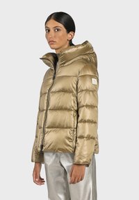 Giacca puffer color oro chiaro con colletto alto, chiusura con zip, polsini a costine e una toppa con logo sulla manica. Texture liscia e lucida.