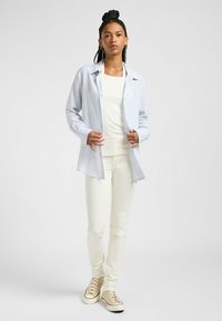 Camisa de botones a rayas azul claro sobre una camiseta blanca de canalé, combinada con pantalones ajustados crema y zapatillas beige. Tela suave y ajuste relajado.