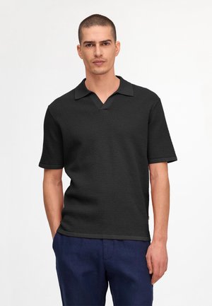 Jeune homme aux cheveux courts portant un polo noir texturé à manches courtes et un pantalon bleu marine, debout devant un fond uni.