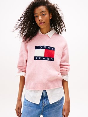 Rosa genser med ribbestrikket kant og mansjetter, med en "TOMMY JEANS" logo i marineblått, hvitt og rødt på forsiden. Brukt over en hvit skjorte med krage.