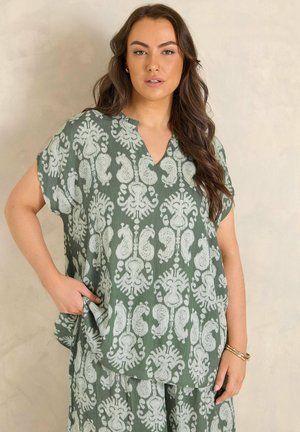 Kvinde med langt brunt hår iført løstsiddende top og matchende bukser med grønne og hvide mønstre, stående med den ene hånd i lommen.