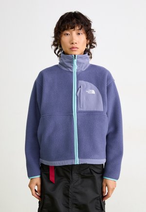 Giacca in pile di colore viola con colletto alto, cerniera azzurro chiaro in contrasto e taschino sul petto con logo. Tessuto morbido e strutturato.