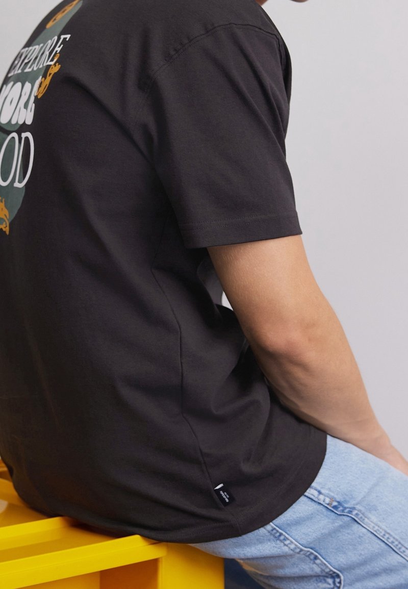 Personne portant un t-shirt graphique noir et un jean bleu clair, assise sur un banc jaune vif devant un fond uni.