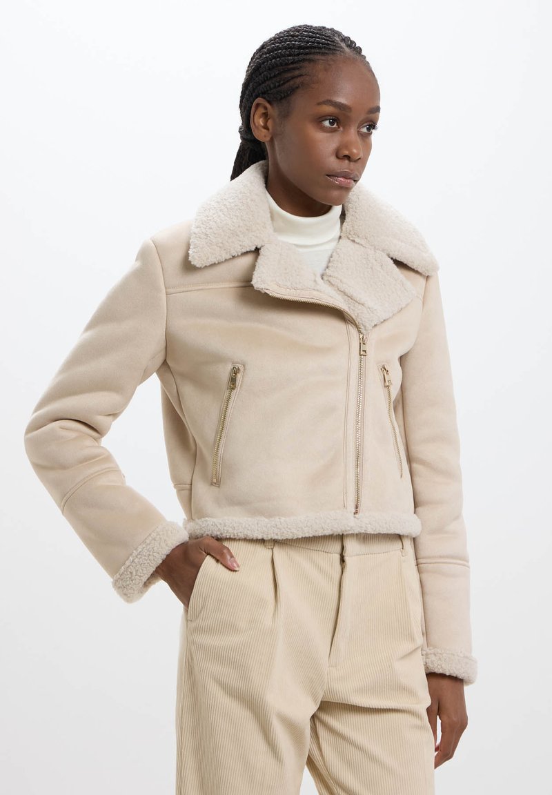 Veste en shearling beige avec un grand col, fermeture éclair avant et poche zippée sur le côté. Tissu texturé avec doublure douce et silhouette cropped.