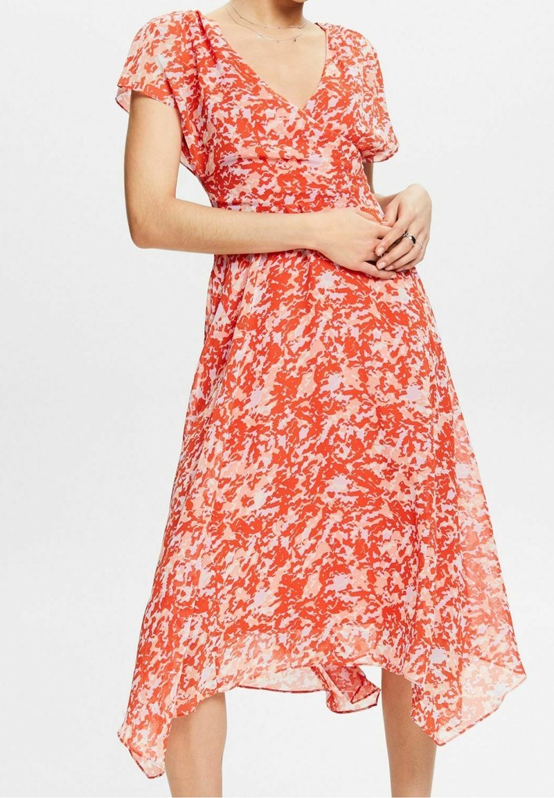 Femme portant une robe à motifs rouges et roses avec un col en V, manches courtes et ourlet asymétrique, les mains jointes légèrement à la taille.