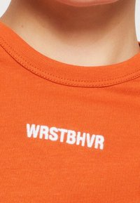 Oranžové žebrované tričko s kulatým výstřihem, na hrudi s bílým vyšitým textem "WRSTBHVR". Měkká, přiléhavá struktura látky.