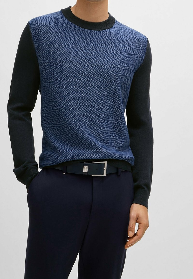 Stickad tröja med ett blått fiskbensmönster på framsidan, mörkblåa långärmar och rund halsringning. Slät textur.