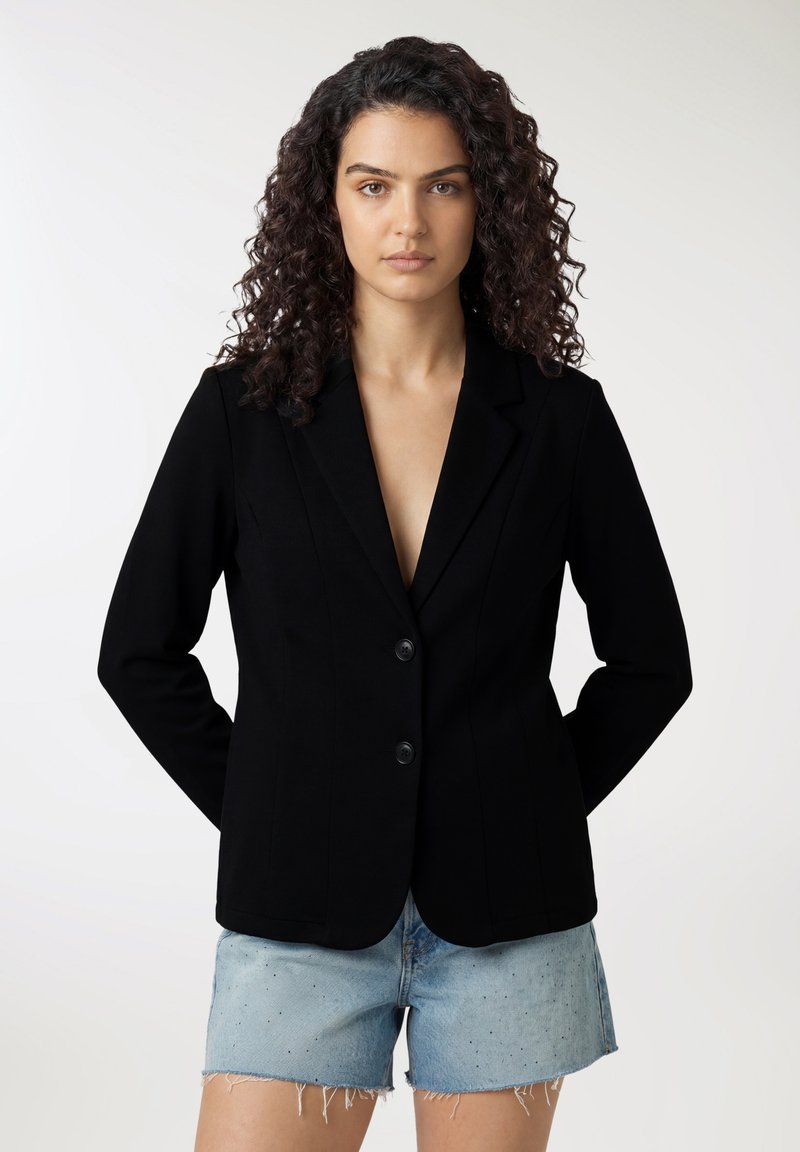 Donna con capelli ricci che indossa un blazer nero con bottoni e pantaloncini di jeans azzurri, in piedi davanti a uno sfondo semplice.