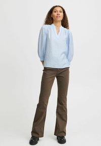 Blusa azzurro chiaro, con texture, scollo a V e maniche a sbuffo 3/4, abbinata a pantaloni marroni svasati e stivali neri, in piedi su uno sfondo neutro.