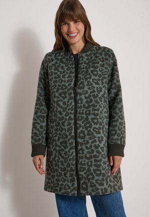 Frau lächelt, trägt eine lange Reißverschlussjacke mit dunkelgrünem und grauem Leopardenmuster und blaue Jeans, steht vor einem schlichten Hintergrund.