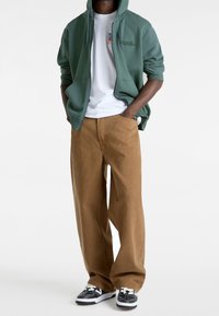 Grön zip-up hoodie över en vit T-shirt, kombinerad med löst sittande bruna byxor och svarta sneakers med vita detaljer.