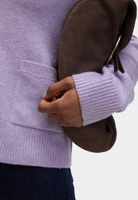 Maglione di lana lilla chiaro con polsini a coste e una tasca, tenuto con una pochette in suede marrone. Primo piano sulla mano e sulla trama del tessuto.