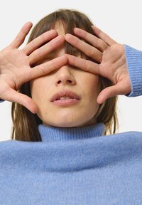 Maglione a collo alto azzurro chiaro con colletto a coste, morbida texture. Mani che coprono il viso, con trucco naturale e capelli castani chiari.