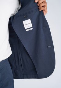 Χέρι που κρατά ανοιχτό ένα μπλέ σκούρο σακάκι, δείχνοντας την εσωτερική ετικέτα με την επιγραφή "Lindbergh Regular Fit Superflex Recycled Fibers" και τις λεπτομέρειες της κουμπότρυπας.