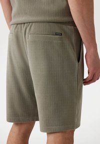 Pantaloni corti grigi con una texture a costine, cintura elastica e tasche posteriori. Presenta una piccola etichetta nera "GUESS" sulla tasca destra.