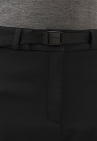 Mustat housut, joissa on sileä pinta, litteä etuosa, sivutasku ja musta vyö suorakaiteen muotoisella soljella.