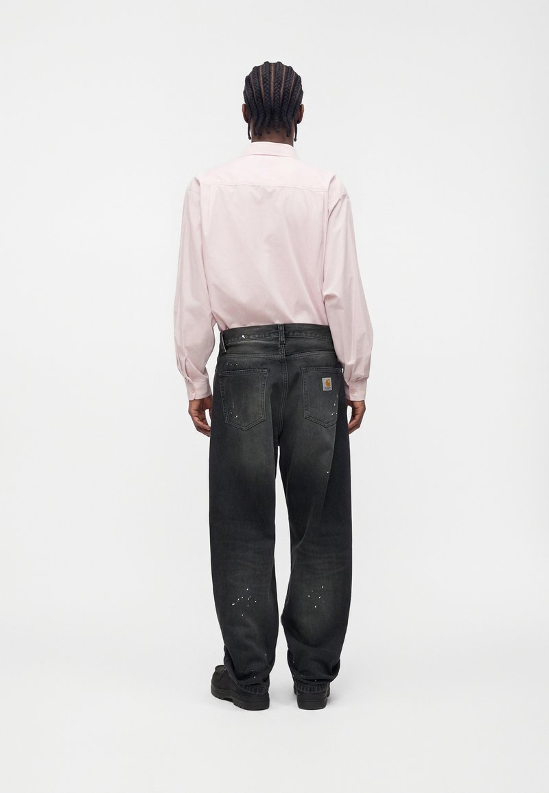 Person with braided hair stands facing away wearing a light pink long-sleeve shirt, faded black jeans, and black shoes on a white background.
