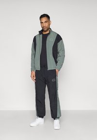 Tumedat roheline ja must treeningkomplekt, millel on elastsed mansetid ja paelalõng. Omab logo aktsente ja kontrastset paneeli disaini.