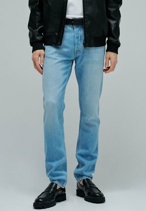 Hombre con jeans azul claro de pierna recta, cinturón negro, chaqueta de cuero negra y mocasines de cuero negros de pie sobre fondo gris.