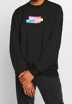 Person iført en sort sweatshirt med et lyserødt og blåt Diesel-logo, armene krydset om livet, kombineret med mørke jeans.