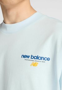 Sudadera de algodón azul claro con cuello redondo, que presenta un logotipo azul de "new balance" y un acento amarillo debajo. Textura suave, diseño ajustado.