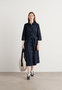 Marineblauwe button-up jurk van katoen, met een kraag, lange mouwen en een voorzak. Combinatie met een beige tas en sneakers.