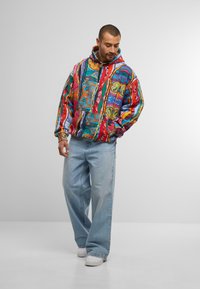 Mann in bunter, gemusterter Kapuzenjacke, hellblauen, weit geschnittenen Jeans und weißen Sneakers, steht mit einer Hand in der Tasche vor einem einfarbigen Hintergrund.