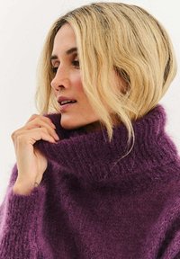Fialový pletený sveter s vysokým rebrovaným golierom, ktorý má jemnú, nadýchanú textúru. Dizajn je voľný, ktoré sa voľne vlní okolo krku.