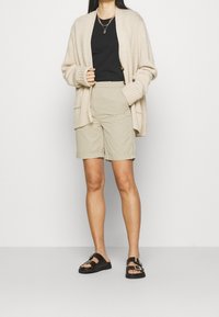 Cardigan lavorato a maglia beige, top nero, pantaloni corti crema con risvolti, e sandali marroni con accenti in oro.