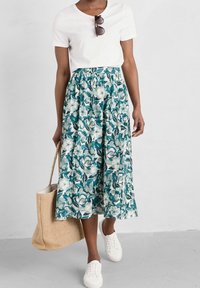 Spódnica midi w kwiatowy wzór w odcieniach zieleni i niebieskiego, z marszczonym pasem. Połączona z białym T-shirtem i białymi trampkami, trzymająca torbę ze słomy.