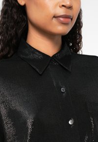Camicia nera con bottoni, caratterizzata da una finitura testurizzata e un colletto classico. Include una tasca sul petto e bottoni neri a contrasto.