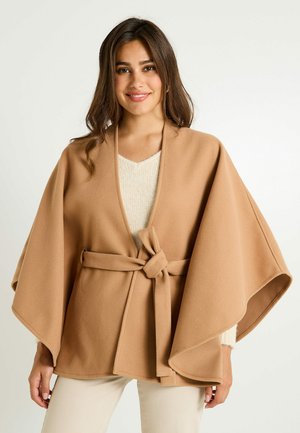 Poncho en polaire beige avec des manches larges et une taille nouée. Présente une texture douce et un tricot crème en V en dessous.