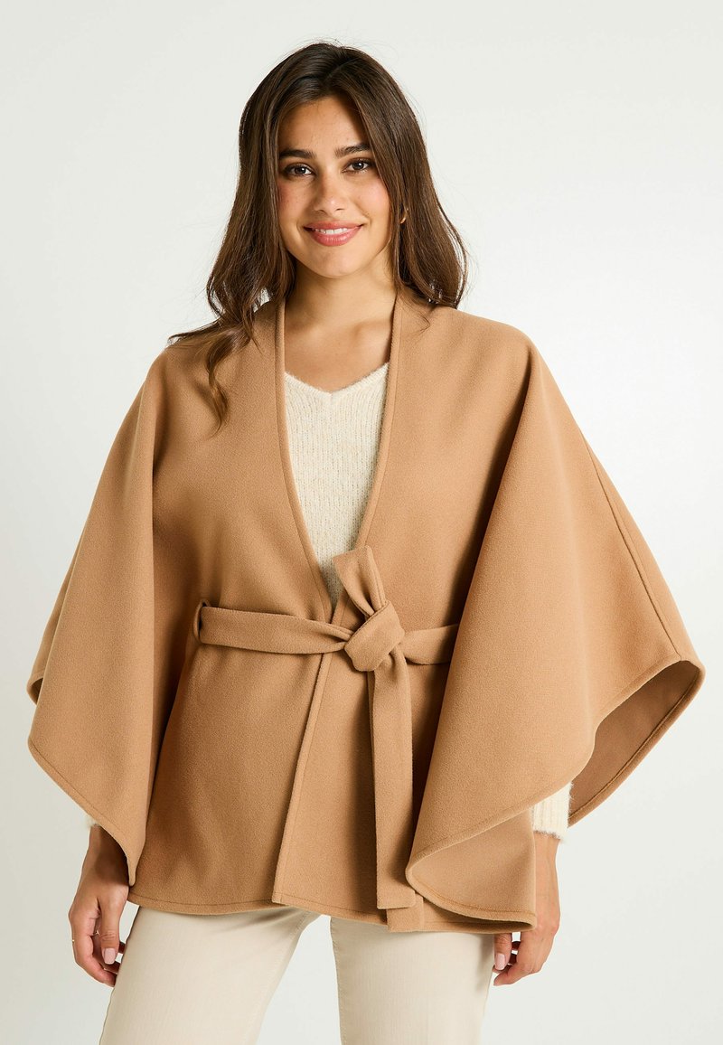 Poncho de forro polar beige con mangas anchas y cintura ajustada. Presenta una textura suave y un jersey de punto crema con escote en V debajo.