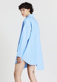 Jeune personne avec des cheveux courts et bouclés portant une chemise oversize bleu clair à rayures, montrant un tatouage d'abeille sur la cuisse supérieure.