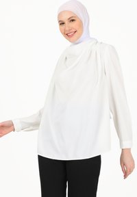 Hvid bluse med draperet stof og lange ærmer. Blød, glat tekstur. Sammen med sorte bukser. Modellen bærer en hvid hijab.