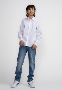 Witte langemouwens blouse met kraag, gecombineerd met lichtblauwe jeans en witte sneakers, staand voor een effen achtergrond.