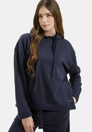 Młoda kobieta o długich brązowych włosach, ubrana w luźną, ciemnogranatową bluzę z kapturem i pasujące spodnie, stoi z jedną ręką w kieszeni.