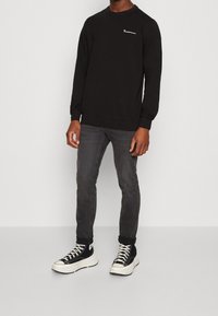 Sweat-shirt noir à col rond, avec un petit logo blanc sur la poitrine. Associé à un jean skinny gris foncé et des baskets noires.