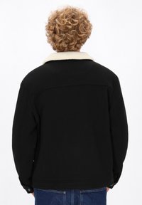 Persona con capelli biondi ricci che indossa una giacca nera con colletto in pile beige e jeans blu, girato di spalle su uno sfondo bianco.