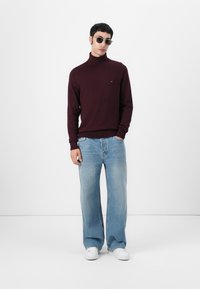 Pull à col roulé bordeaux, coupe décontractée, associé à un jean bleu clair à jambes larges et des baskets blanches. Les accessoires comprennent des lunettes de soleil grises.