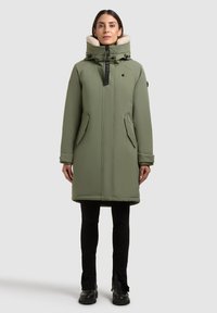 Olivovo zelený prešívaný kabát s vysokým golierom a béžovým umelým kožuchom. Obsahuje zipsové vrecká a predné zapínanie na zips.