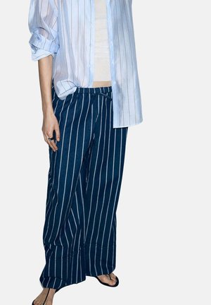 Persona que lleva pantalones sueltos azul oscuro con rayas blancas verticales, una camisa de rayas azul claro con mangas remangadas, chanclas y un anillo en la mano izquierda.