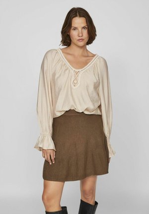 Blusa de manga larga color beige claro con detalle de encaje en el escote, combinada con una falda de punto marrón. Modelo lleva botas negras hasta la rodilla.