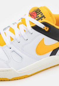 Vit sneaker med gula accenter, svart häl och vita snören. Har perforerade sektioner och en gummisula med gula detaljer.