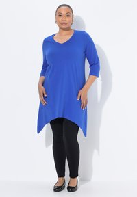 Blaue Tunika mit V-Ausschnitt, Dreiviertelärmeln und asymmetrischem Saum. Kombiniert mit schwarzen Leggings und flachen Schuhen. Glatte Textur.