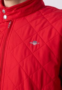 Giacca trapuntata rossa con colletto alto, bottoni a pressione argentati e logo ricamato sul petto sinistro. Texture del tessuto liscia.